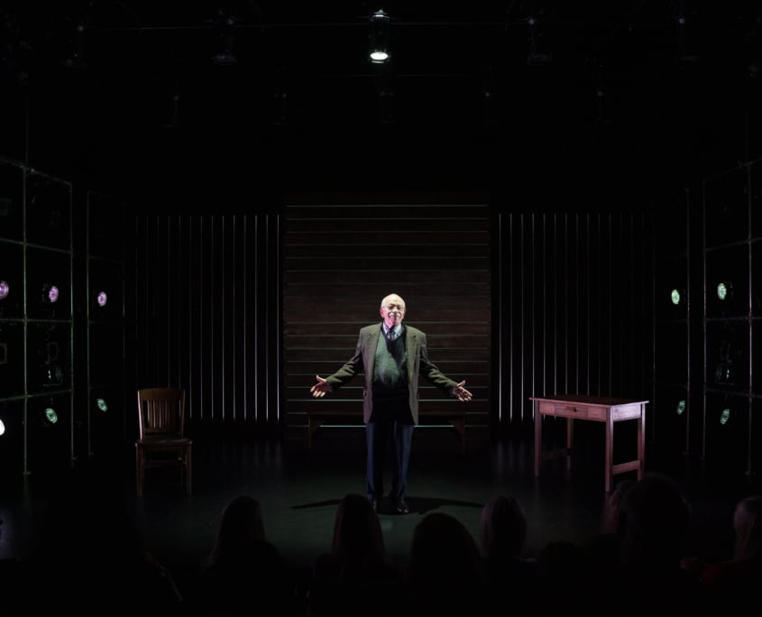 Actor Kenneth Tigar, playing Holocaust survivor Eddie Jaku, stands on a stage under dramatic lighting with shadows cast around them, addressing an unseen crowd.