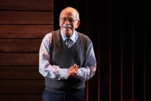 Actor Kenneth Tigar, playing Holocaust survivor Eddie Jaku, smiles out at the audience with his hands clasped in front of him, suggesting a moment of connection or gratitude.