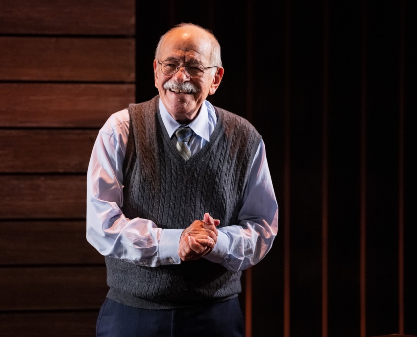 Actor Kenneth Tigar, playing Holocaust survivor Eddie Jaku, smiles out at the audience with his hands clasped in front of him, suggesting a moment of connection or gratitude.