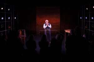 Actor Kenneth Tigar, playing Holocaust survivor Eddie Jaku, stands on a stage lit by spotlights, facing an audience. Wooden elements, a chair, and a table frame the scene.
