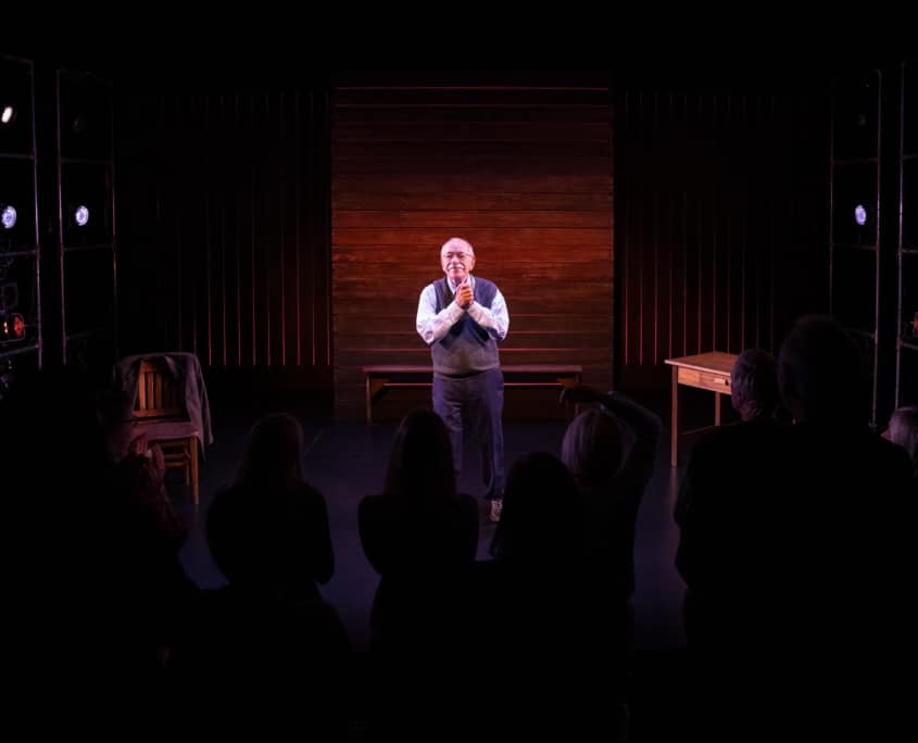 Actor Kenneth Tigar, playing Holocaust survivor Eddie Jaku, stands on a stage lit by spotlights, facing an audience. Wooden elements, a chair, and a table frame the scene.