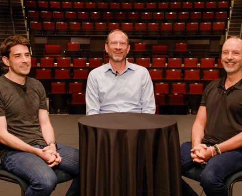 Three individuals are seated around a small round table covered with a black cloth. They are positioned in front of an empty auditorium with rows of red seats. The person on the left is wearing a dark gray t-shirt and jeans, the person in the middle is wearing a light blue button-up shirt and glasses, and the person on the right is wearing a black polo shirt and jeans.