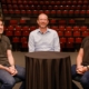 Three individuals are seated around a small round table covered with a black cloth. They are positioned in front of an empty auditorium with rows of red seats. The person on the left is wearing a dark gray t-shirt and jeans, the person in the middle is wearing a light blue button-up shirt and glasses, and the person on the right is wearing a black polo shirt and jeans.