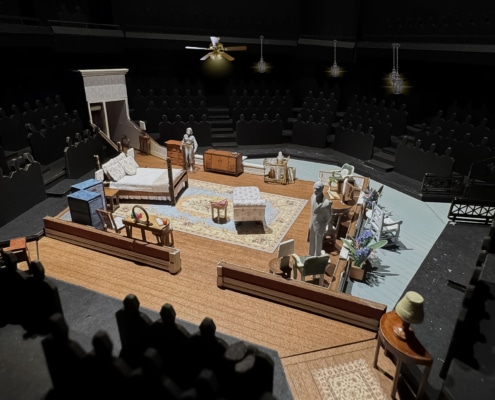 A set model of Cat on a Hot Tin Roof showing a bedroom in a Southern plantation with a bed, vanity, chaise lounge, rug, various chairs, and openings into a hallway and onto a veranda.