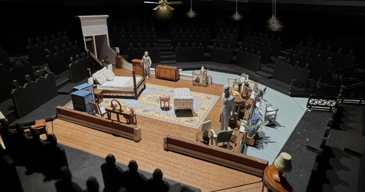 A set model of Cat on a Hot Tin Roof showing a bedroom in a Southern plantation with a bed, vanity, chaise lounge, rug, various chairs, and openings into a hallway and onto a veranda.