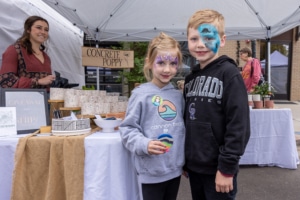 Two children with face paint stand in front of a booth at an outdoor market. The booth displays a sign reading 'CONCRETE POPPY' and various items for sale, with a smiling vendor behind the table.