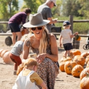 A person wearing a hat and sunglasses kneels on the ground, smiling at a small child who is picking up a pumpkin. Several pumpkins are scattered around them, with other people in the background also selecting pumpkins.