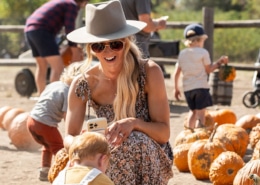 A person wearing a hat and sunglasses kneels on the ground, smiling at a small child who is picking up a pumpkin. Several pumpkins are scattered around them, with other people in the background also selecting pumpkins.