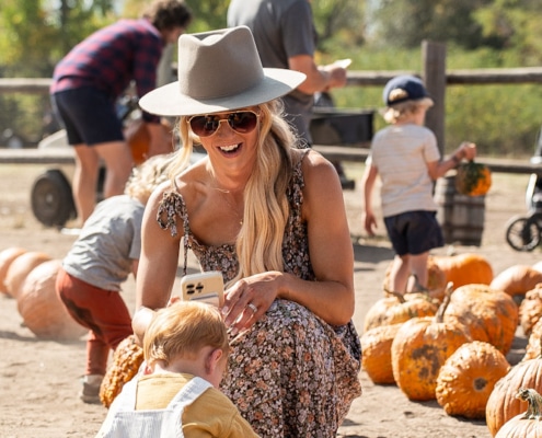 A person wearing a hat and sunglasses kneels on the ground, smiling at a small child who is picking up a pumpkin. Several pumpkins are scattered around them, with other people in the background also selecting pumpkins.