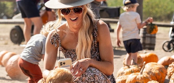 A person wearing a hat and sunglasses kneels on the ground, smiling at a small child who is picking up a pumpkin. Several pumpkins are scattered around them, with other people in the background also selecting pumpkins.
