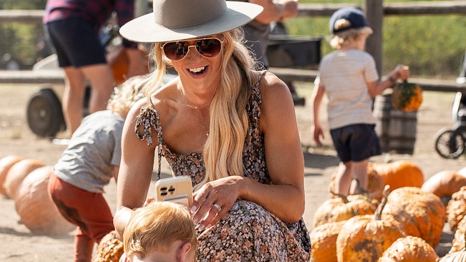 A person wearing a hat and sunglasses kneels on the ground, smiling at a small child who is picking up a pumpkin. Several pumpkins are scattered around them, with other people in the background also selecting pumpkins.