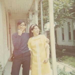 Two people standing on a porch, one wearing glasses and a dark sweater, the other in a yellow dress with white trim. The person in the yellow dress has an arm around the other person. The background shows part of a house and some greenery.