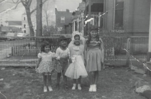 A black and white photograph of four children standing in a yard. The children are dressed in various outfits, with one child wearing a dress and veil, suggesting a special occasion. The background shows a fence, trees, and houses.