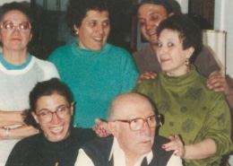 Six people posing closely together indoors. An older individual wearing glasses and a dark vest over a white shirt is seated in front. To the left, a person with glasses wears a black shirt with yellow text. Four others stand behind them, smiling and interacting. The background includes furniture and interior walls.