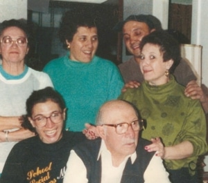 Six people posing closely together indoors. An older individual wearing glasses and a dark vest over a white shirt is seated in front. To the left, a person with glasses wears a black shirt with yellow text. Four others stand behind them, smiling and interacting. The background includes furniture and interior walls.