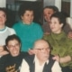Six people posing closely together indoors. An older individual wearing glasses and a dark vest over a white shirt is seated in front. To the left, a person with glasses wears a black shirt with yellow text. Four others stand behind them, smiling and interacting. The background includes furniture and interior walls.