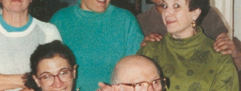 Six people posing closely together indoors. An older individual wearing glasses and a dark vest over a white shirt is seated in front. To the left, a person with glasses wears a black shirt with yellow text. Four others stand behind them, smiling and interacting. The background includes furniture and interior walls.