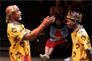 An older Black man knights a younger Black man with a large, slotted spoon, another Black man watches the scene, smiling. The young man is wearing a gold crown, all the men are wearing yellow and orange Hawiian shirts with flaming hearts on them.