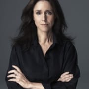 Portrait of Julie Taymor with long dark hair, wearing a black shirt, standing against a gray background with arms crossed.