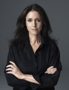 Portrait of Julie Taymor with long dark hair, wearing a black shirt, standing against a gray background with arms crossed.