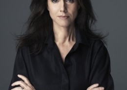 Portrait of Julie Taymor with long dark hair, wearing a black shirt, standing against a gray background with arms crossed.