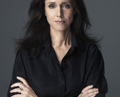 Portrait of Julie Taymor with long dark hair, wearing a black shirt, standing against a gray background with arms crossed.