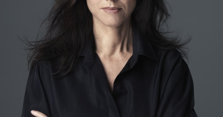 Portrait of Julie Taymor with long dark hair, wearing a black shirt, standing against a gray background with arms crossed.