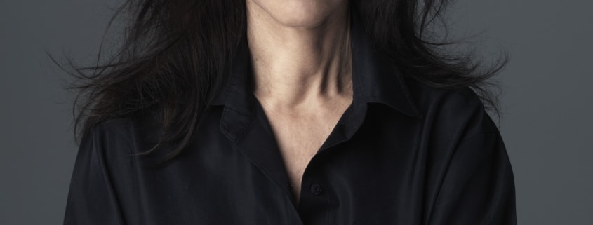 Portrait of Julie Taymor with long dark hair, wearing a black shirt, standing against a gray background with arms crossed.