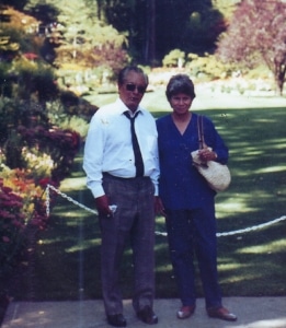 Two individuals standing on a paved path in a garden or park, surrounded by lush greenery, colorful flowers, and trees. One wears a white shirt with a dark tie and gray pants, holding an object; the other wears blue attire and carries a woven handbag. A white chain barrier is visible behind them.