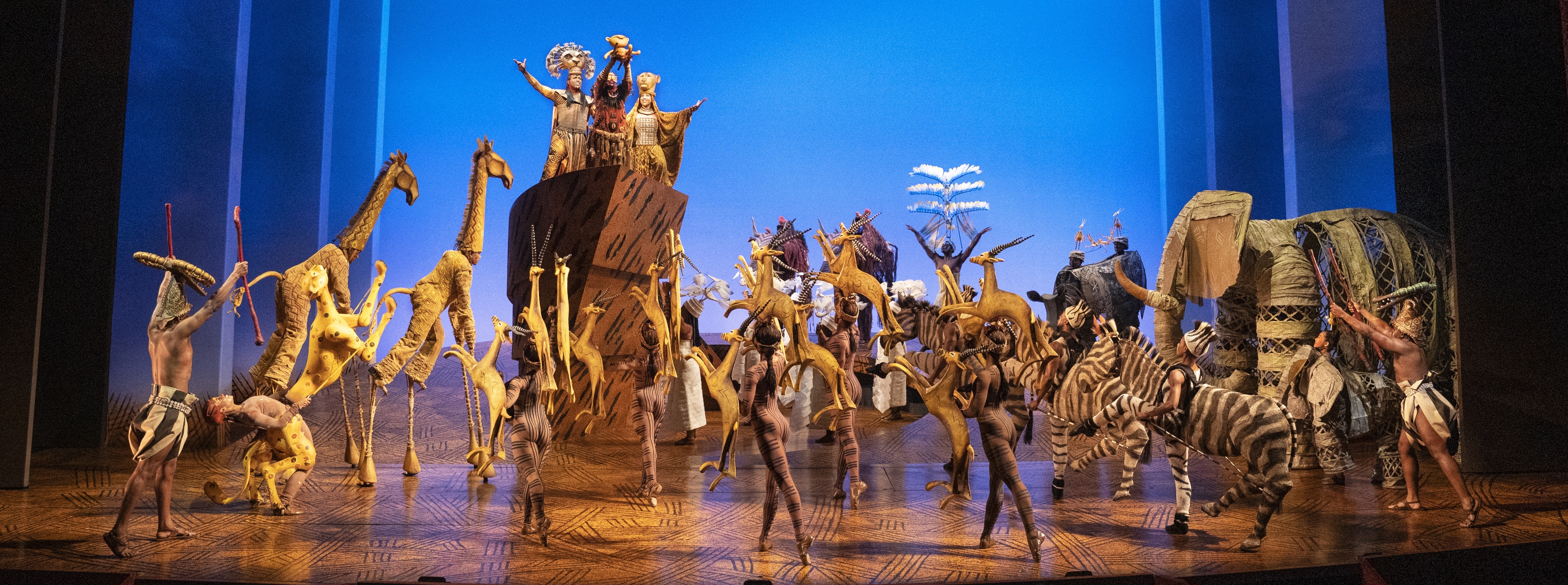 A vibrant stage performance featuring actors in elaborate animal costumes, including giraffes, zebras, and elephants, set against a bright blue backdrop with stylized clouds