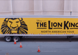 Photo of a semitruck on a street outside a theatre what has the Lion King logo (illustration of a lion head on a yellow background and the title of the show) painted on the side