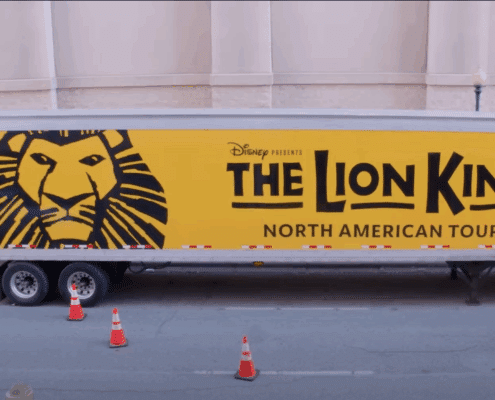 Photo of a semitruck on a street outside a theatre what has the Lion King logo (illustration of a lion head on a yellow background and the title of the show) painted on the side