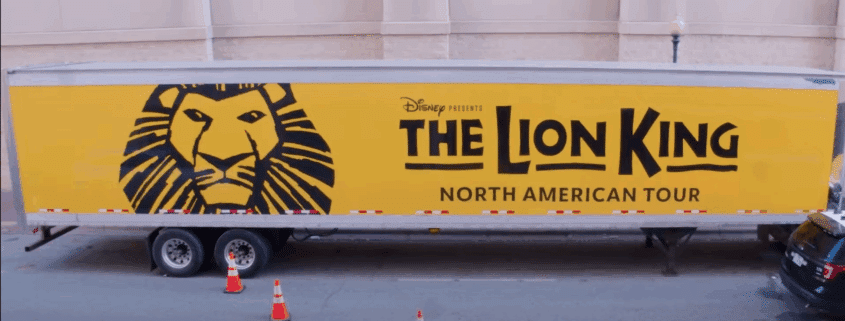 Photo of a semitruck on a street outside a theatre what has the Lion King logo (illustration of a lion head on a yellow background and the title of the show) painted on the side