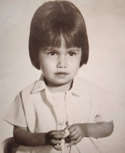 Black and white photograph of a young child with short hair and bangs, wearing a light-colored shirt and holding a small object in both hands. The background is plain, emphasizing the child's face and upper body.