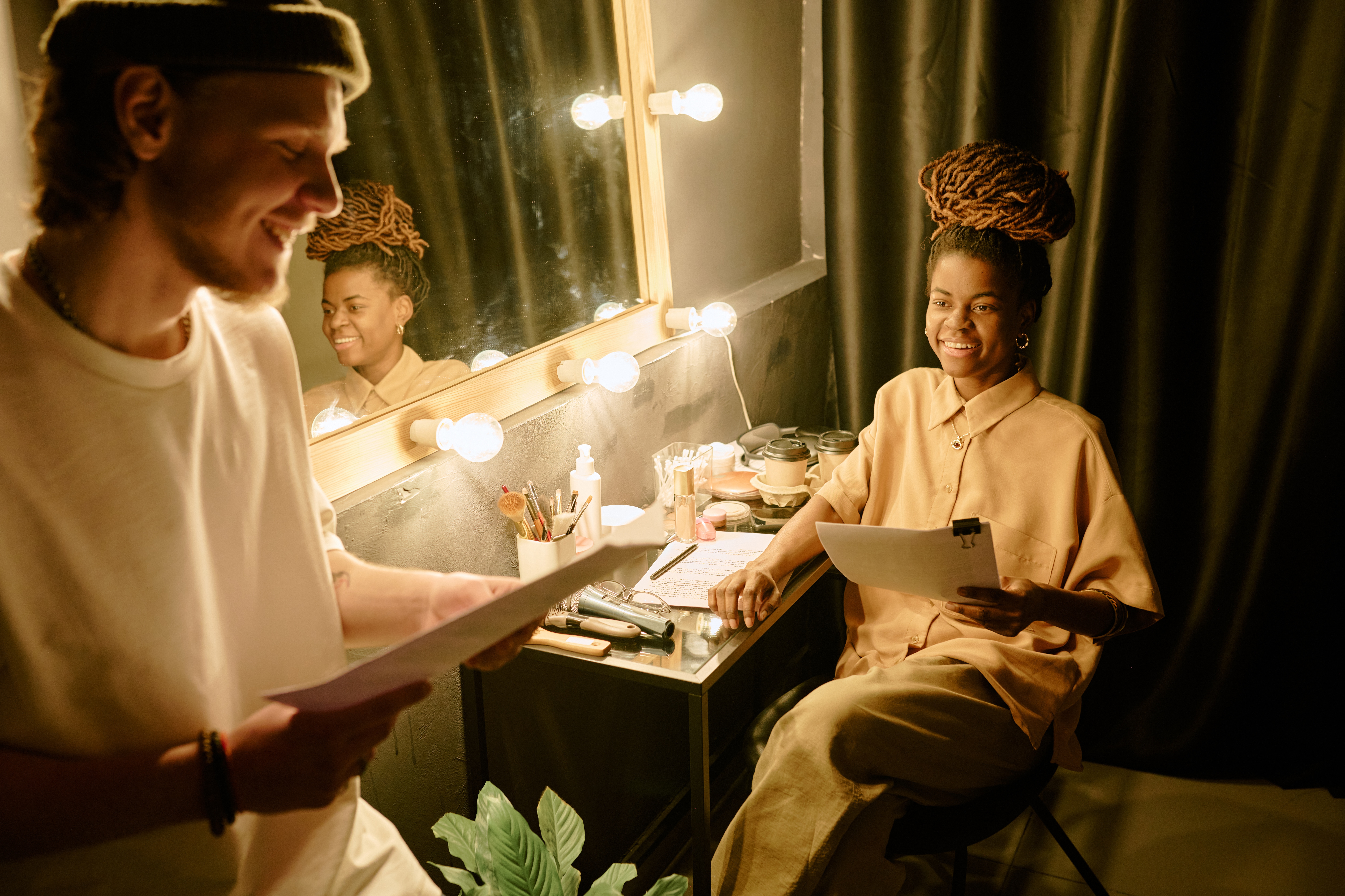 Two actors in a dressing room in front of a mirror surrounded by lights. On the left is a young man in knit cap and t-shirt holding a script. On the right is a young lady seated in neutral-colored clothing, smiling with a script in her hand.