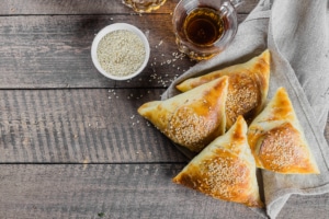 Four triangular meat-filled samosas topped with sesame seeds, served on a cloth with a bowl of sesame seeds and a glass of tea on a wooden surface.