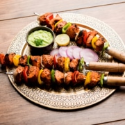 Grilled chicken tikka skewers with bell peppers and onions, served on a decorative plate with green sauce, lemon slice, and red onion, on a rustic wooden table.
