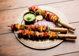 Grilled chicken tikka skewers with bell peppers and onions, served on a decorative plate with green sauce, lemon slice, and red onion, on a rustic wooden table.