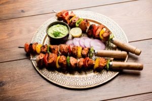 Grilled chicken tikka skewers with bell peppers and onions, served on a decorative plate with green sauce, lemon slice, and red onion, on a rustic wooden table.