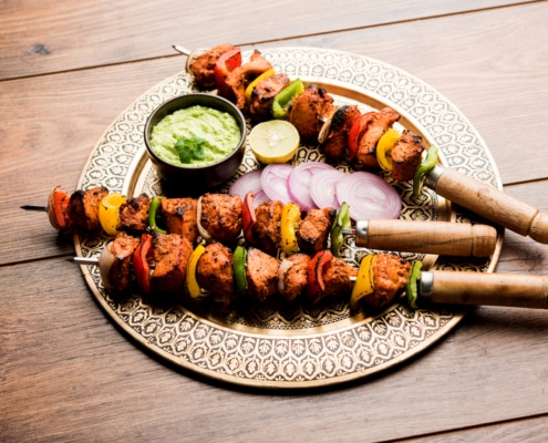 Grilled chicken tikka skewers with bell peppers and onions, served on a decorative plate with green sauce, lemon slice, and red onion, on a rustic wooden table.