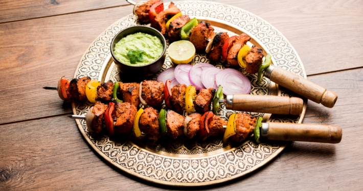 Grilled chicken tikka skewers with bell peppers and onions, served on a decorative plate with green sauce, lemon slice, and red onion, on a rustic wooden table.