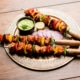 Grilled chicken tikka skewers with bell peppers and onions, served on a decorative plate with green sauce, lemon slice, and red onion, on a rustic wooden table.