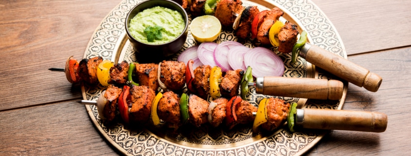 Grilled chicken tikka skewers with bell peppers and onions, served on a decorative plate with green sauce, lemon slice, and red onion, on a rustic wooden table.