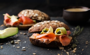 Sliced bagel with lox, avocado slices, and capers, served on a dark surface with crumbs and a bowl of sauce in the background.