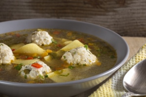 Bowl of clear golden broth soup with meatballs, potato slices, red bell peppers, and herbs, served on a wooden surface with a yellow napkin and spoon.