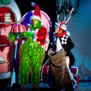 The Grinch, dressed in a green furry costume with a red Santa hat, holds a large pink and white striped cake on a platter. Next to the Grinch is another character dressed as Max the dog, wearing antlers and holding a sack. They are standing in front of colorful cartoon-like scenery from Whoville.