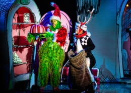 The Grinch, dressed in a green furry costume with a red Santa hat, holds a large pink and white striped cake on a platter. Next to the Grinch is another character dressed as Max the dog, wearing antlers and holding a sack. They are standing in front of colorful cartoon-like scenery from Whoville.