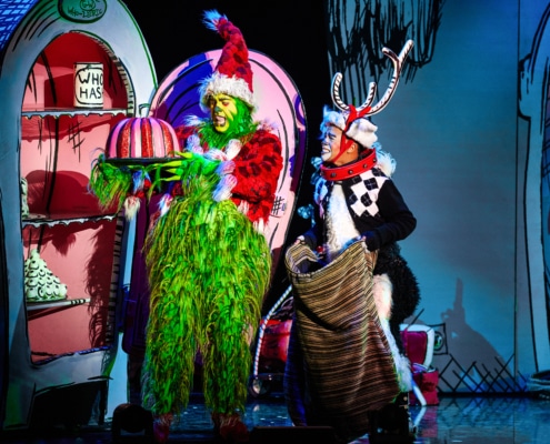 The Grinch, dressed in a green furry costume with a red Santa hat, holds a large pink and white striped cake on a platter. Next to the Grinch is another character dressed as Max the dog, wearing antlers and holding a sack. They are standing in front of colorful cartoon-like scenery from Whoville.
