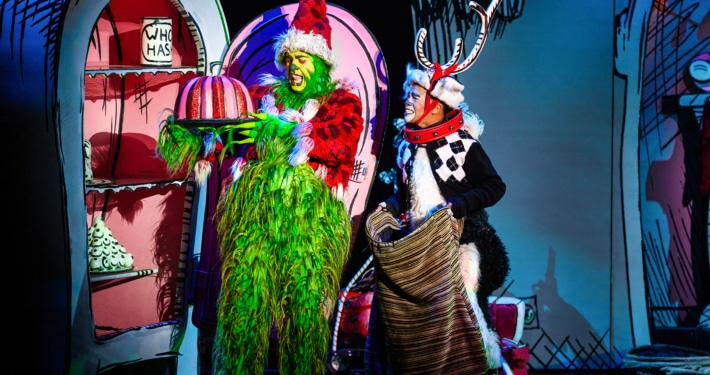 The Grinch, dressed in a green furry costume with a red Santa hat, holds a large pink and white striped cake on a platter. Next to the Grinch is another character dressed as Max the dog, wearing antlers and holding a sack. They are standing in front of colorful cartoon-like scenery from Whoville.