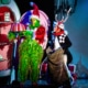 The Grinch, dressed in a green furry costume with a red Santa hat, holds a large pink and white striped cake on a platter. Next to the Grinch is another character dressed as Max the dog, wearing antlers and holding a sack. They are standing in front of colorful cartoon-like scenery from Whoville.